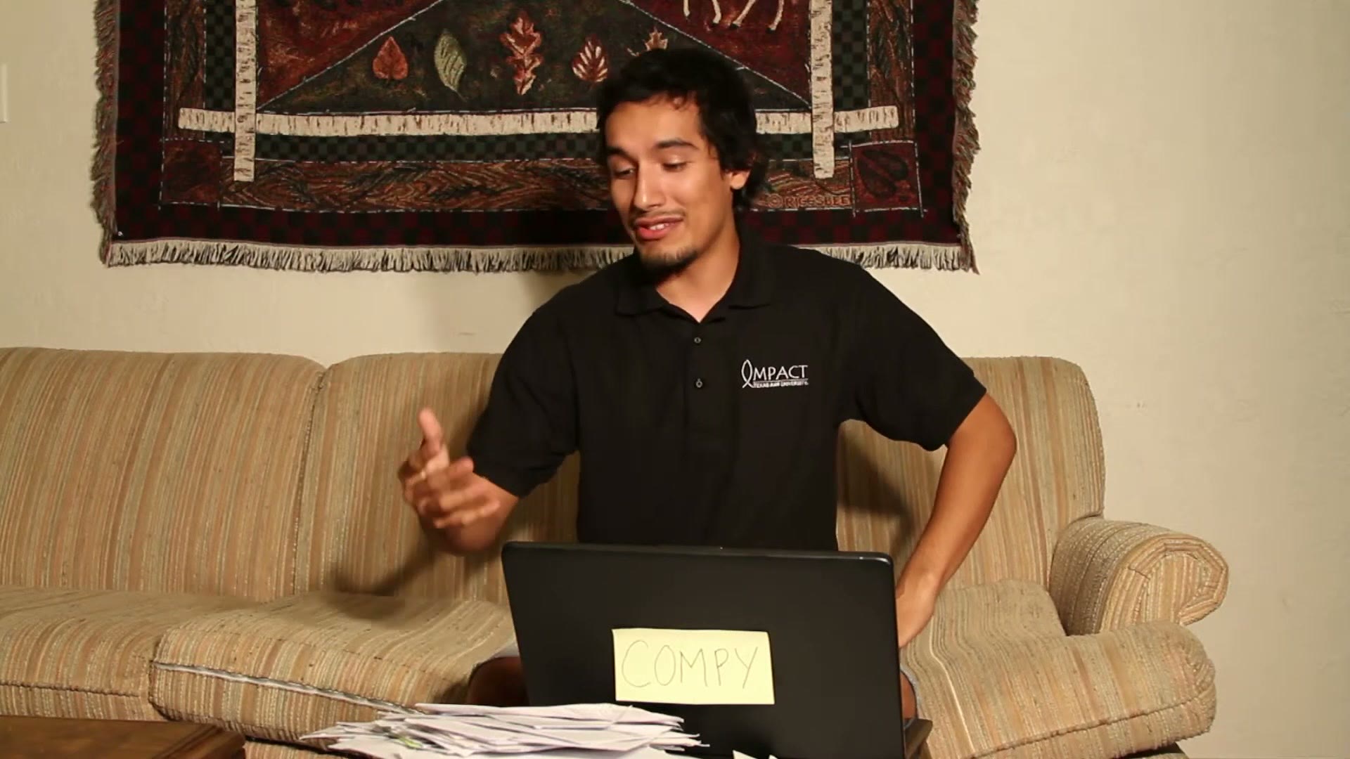 A college student in a black Impact polo sitting on a couch doing paperwork, with a stack of envelopes on the coffee table and a laptop labelled with a handwritten sign