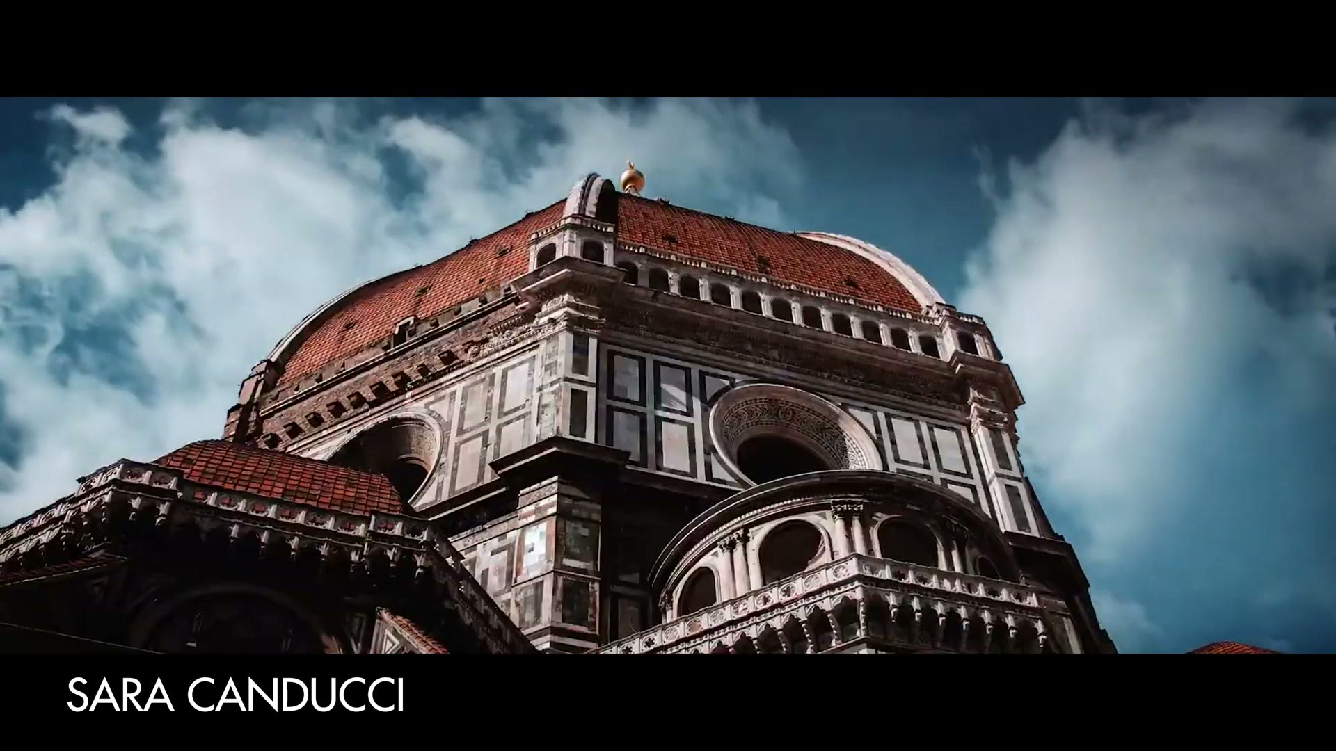 A low angle shot of the dome of the Duomo in Florence against a deep blue sky with clouds, credited on screen to Sara Canducci