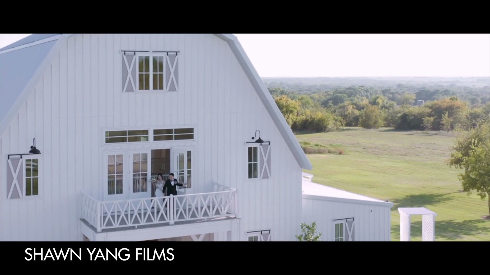An aerial shot of a bride and groom standing on the second floor balcony of a white barn wedding venue with open countryside behind them, credited on screen to Shawn Yang Films