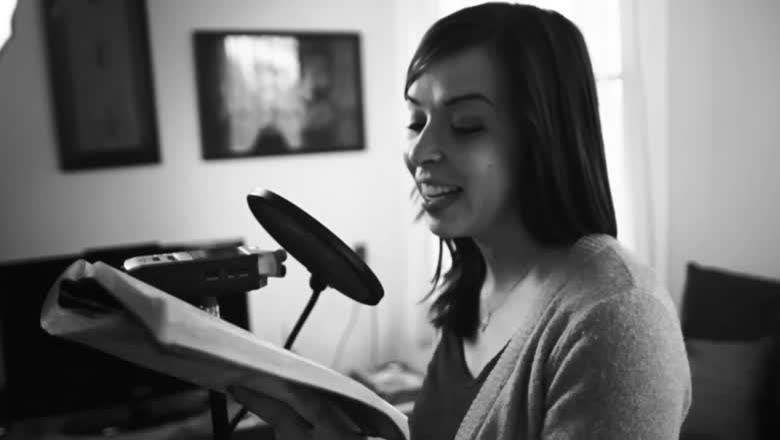 A woman in black and white reading from a page into a microphone with a pop filter, seated in a living room