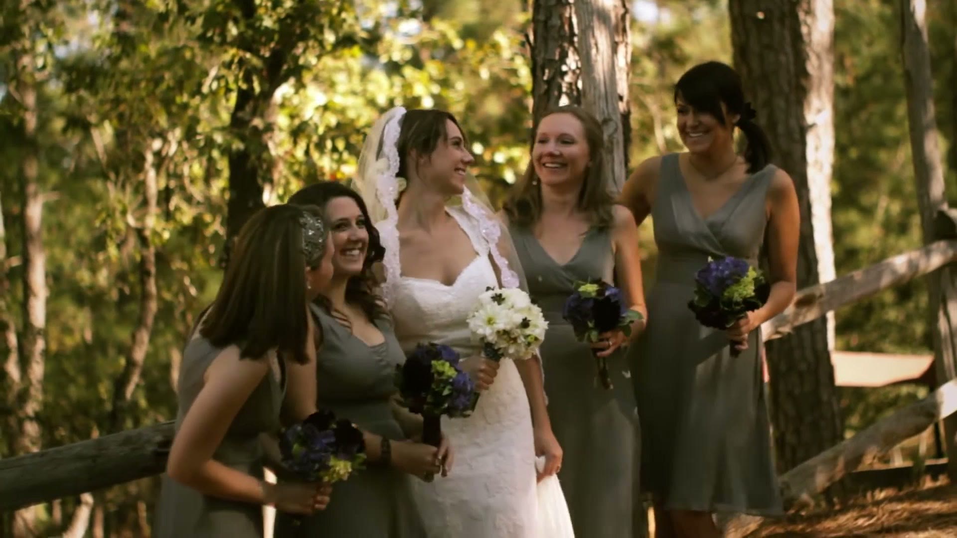 A bride in a lace gown standing with four bridesmaids in sage green dresses among tall pine trees, all holding bouquets of white flowers and dark berries