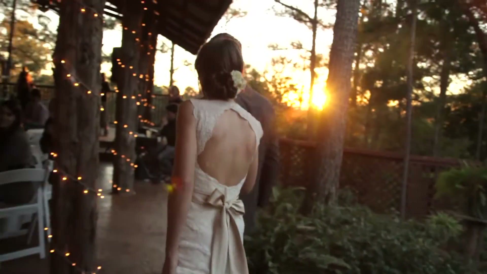 A bride photographed from behind under an open pavilion strung with fairy lights, with the sun setting through pine trees beyond her