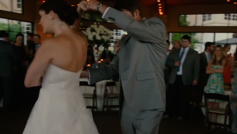 The groom spinning the bride out during their first dance in a glass-walled reception room, guests standing around the floor