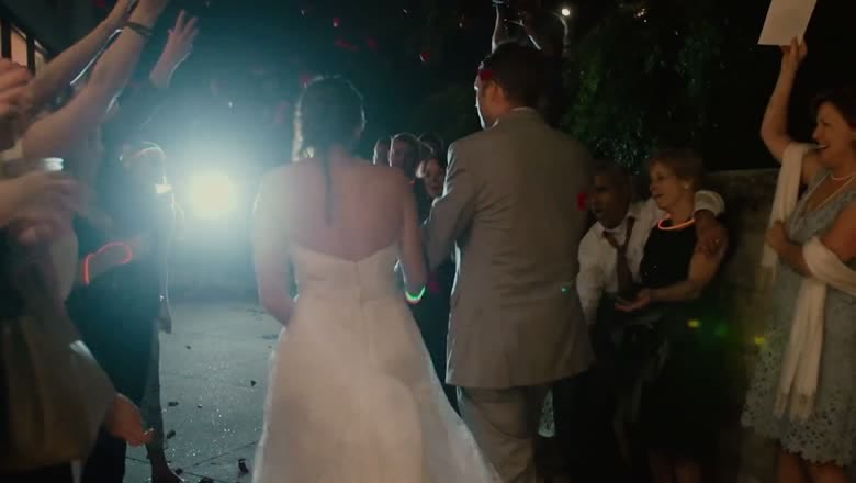 The couple leaving at night through two rows of guests waving glow sticks, lit from behind