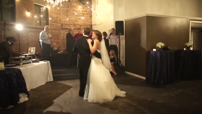 The bride and groom having their first dance in a brick-walled room strung with lights, guests watching from the edges