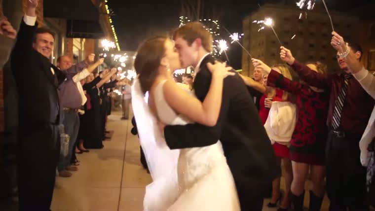The couple kissing on a downtown sidewalk at night between two rows of guests holding sparklers overhead
