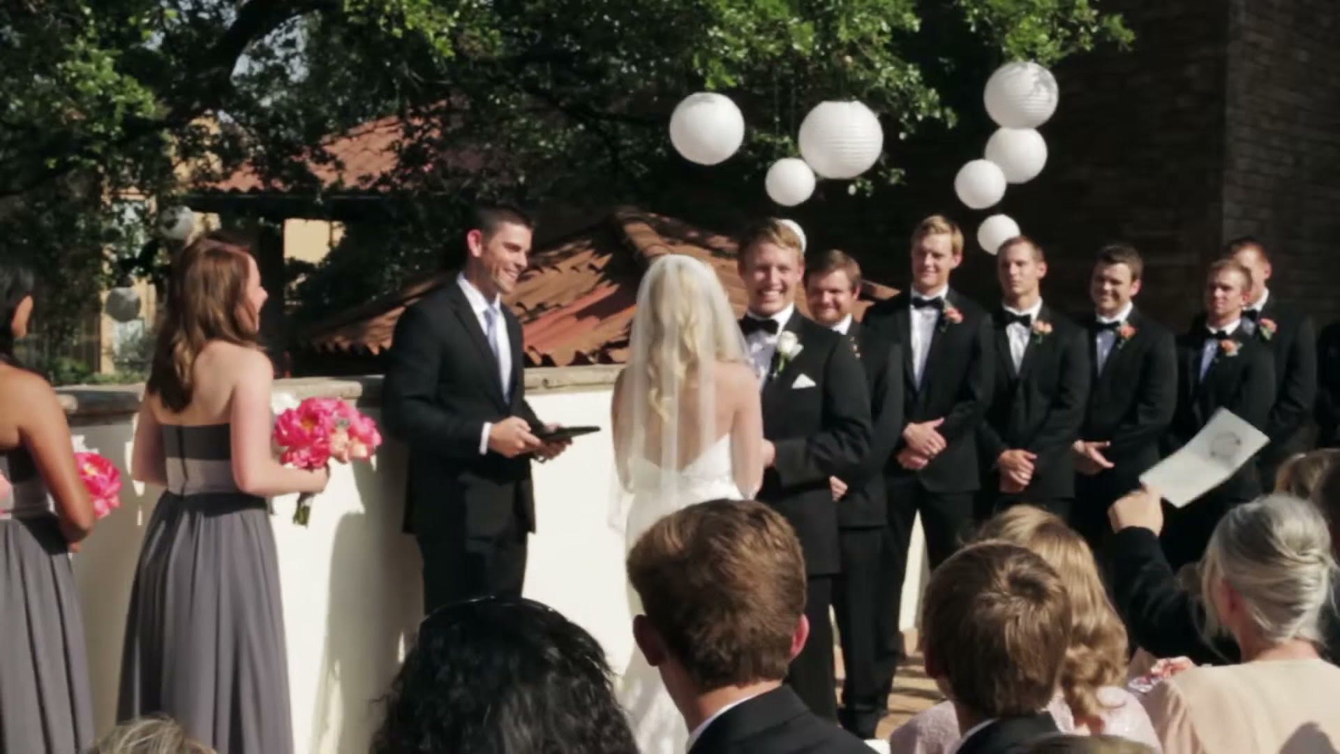 The outdoor ceremony from behind the guests, bride and groom facing the officiant under white paper lanterns hanging from a tree