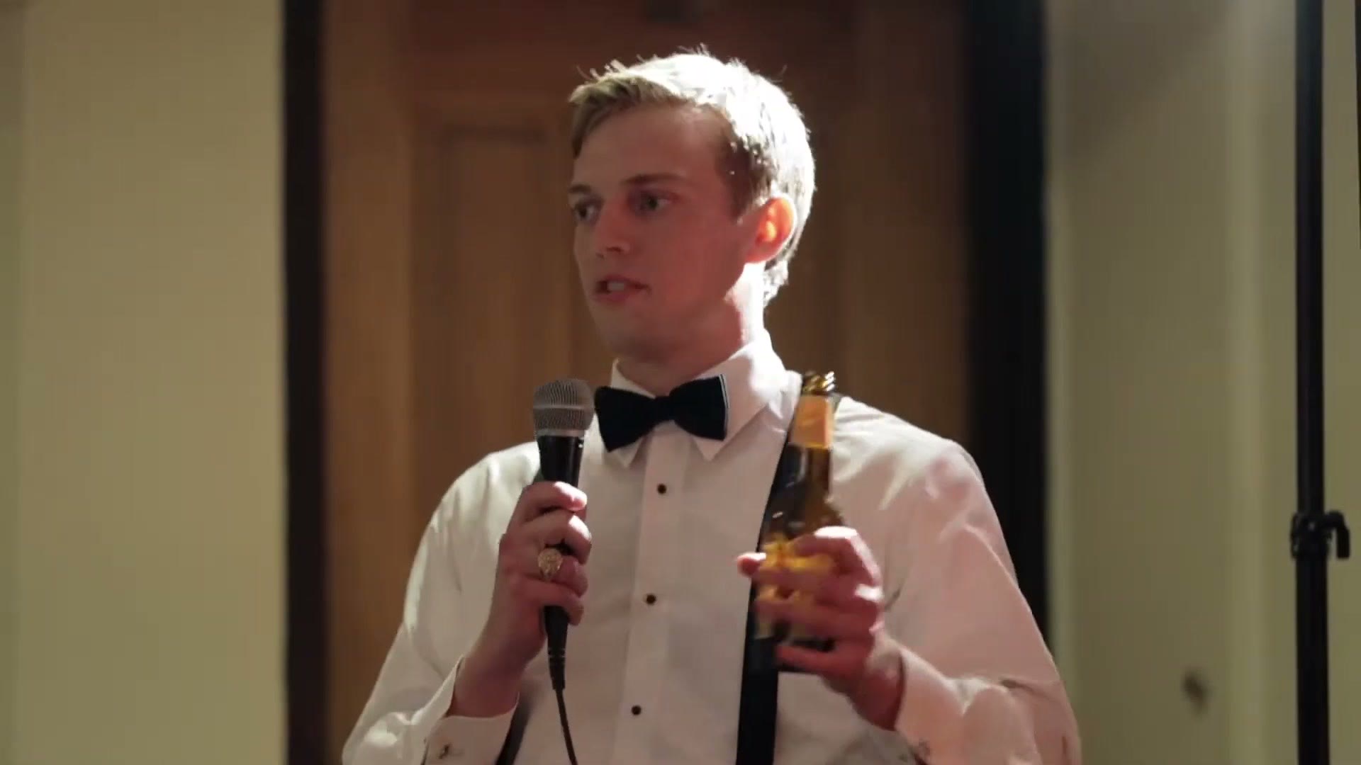 A groomsman giving a toast into a microphone at the reception, beer bottle in his other hand