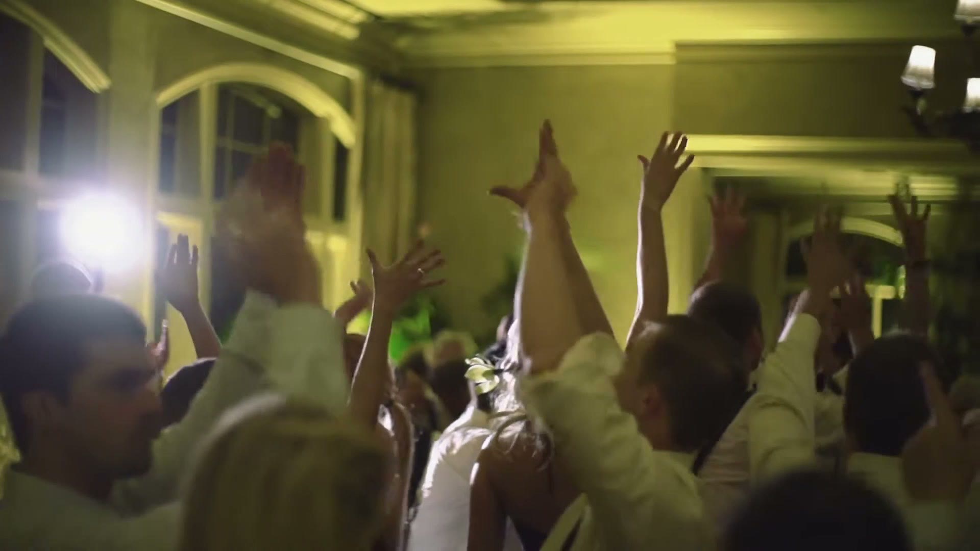 A packed wedding reception dance floor with dozens of guests and their hands in the air under warm light