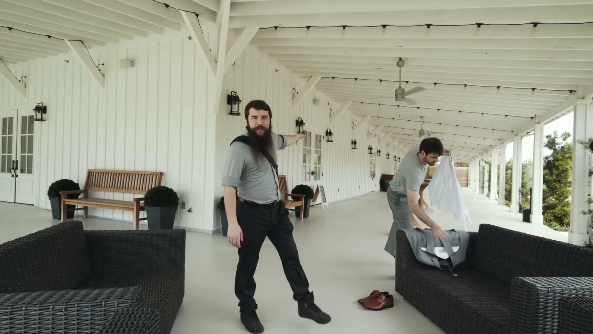 Matt standing on a long covered white porch strung with cafe lights, camera bag over his shoulder, while the groom's party moves out of the dark prep room.