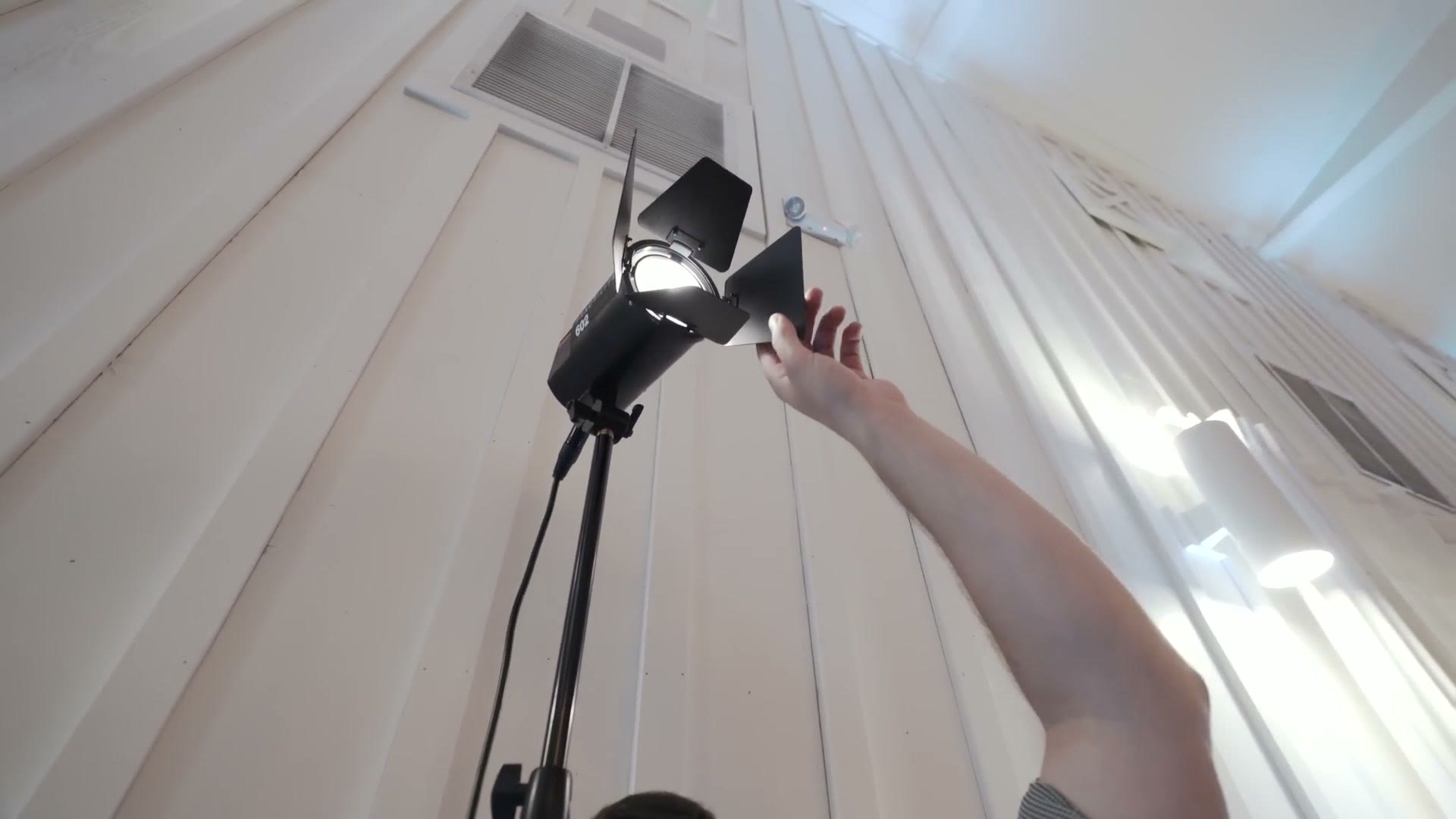 A hand adjusting the barn doors on a small LED light mounted on a stand against a white draped wall.
