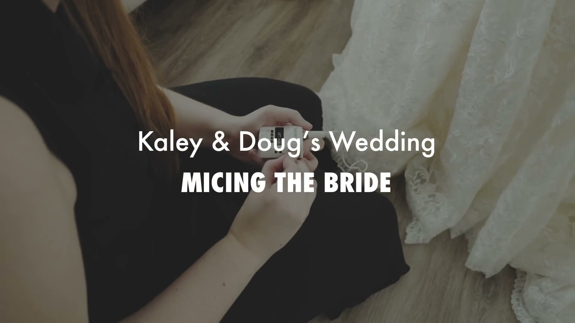 A hand holding a small white lav recorder up against the lace bodice of a hanging wedding dress, with the on-screen title Kaley and Doug's Wedding, Micing The Bride.