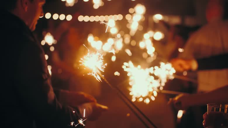 Close on a cluster of lit sparklers throwing sparks in the dark, with string lights out of focus behind