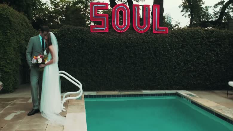 The bride and groom standing together beside a swimming pool with a large red neon SOUL sign mounted on the hedge behind them