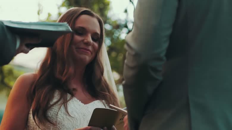 The bride smiling as the groom reads from a small book of vows, the oak tree canopy out of focus behind them
