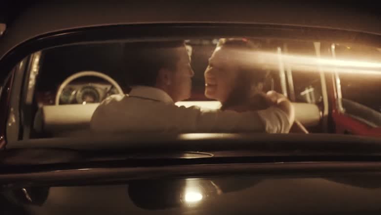 The couple sitting close together inside a vintage car at night, lit warm from outside