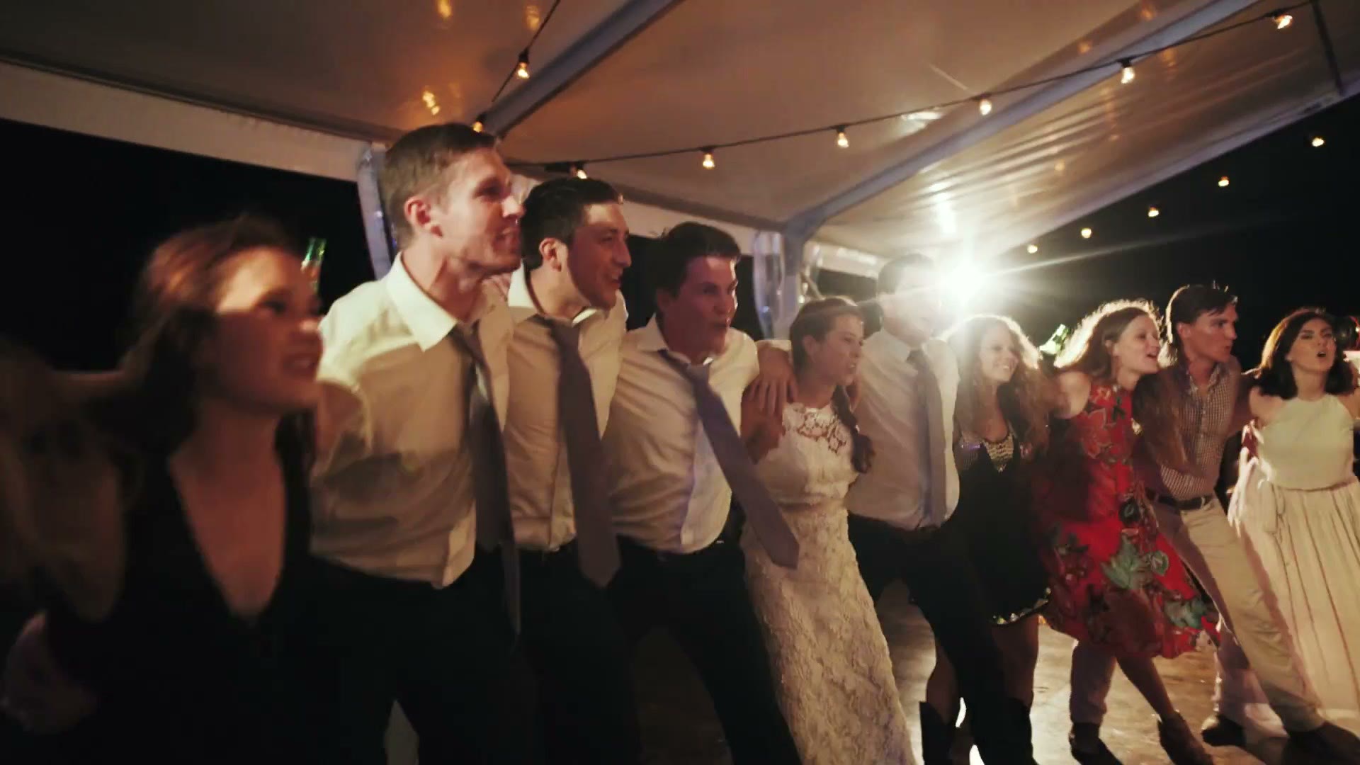 Wedding guests and the bride in a long line with arms around each other, dancing under a marquee strung with festoon lights