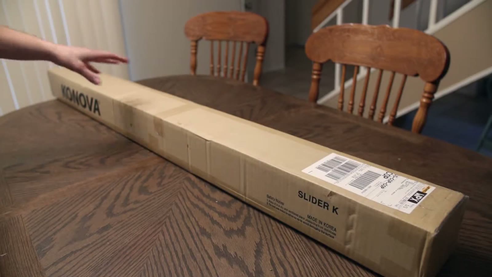 Hand reaching toward a long cardboard Konova Slider K box on a dining table before opening it