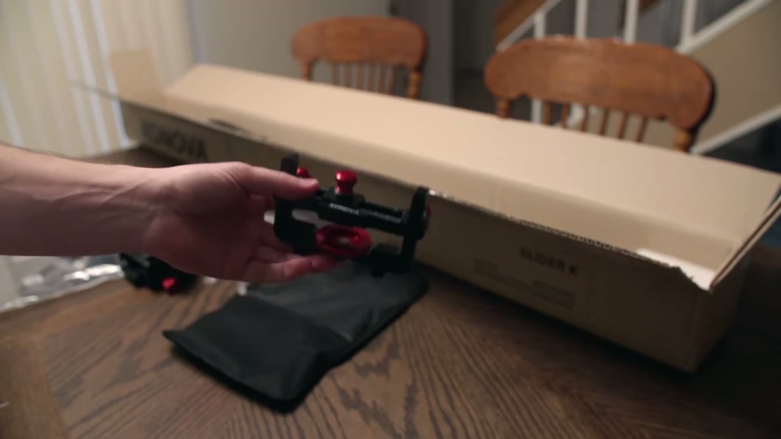 Hand holding the assembled Konova mounting bracket with multiple red knobs beside its black carrying pouch