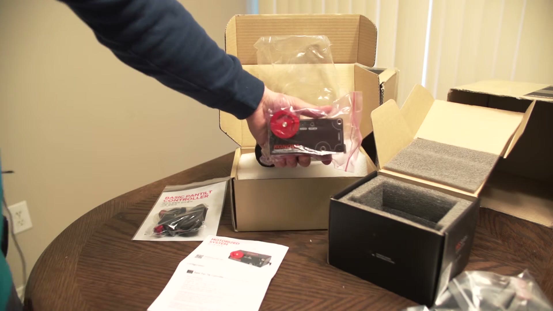 A hand holding the new Konova MS Series motor in its bag, with the motorized system and pan tilt controller manuals on the table below