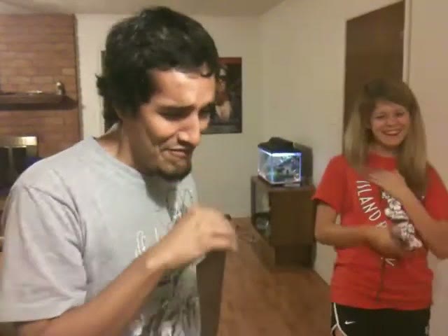A young man grimacing hard after eating something, with a friend laughing at him in the background of an apartment living room