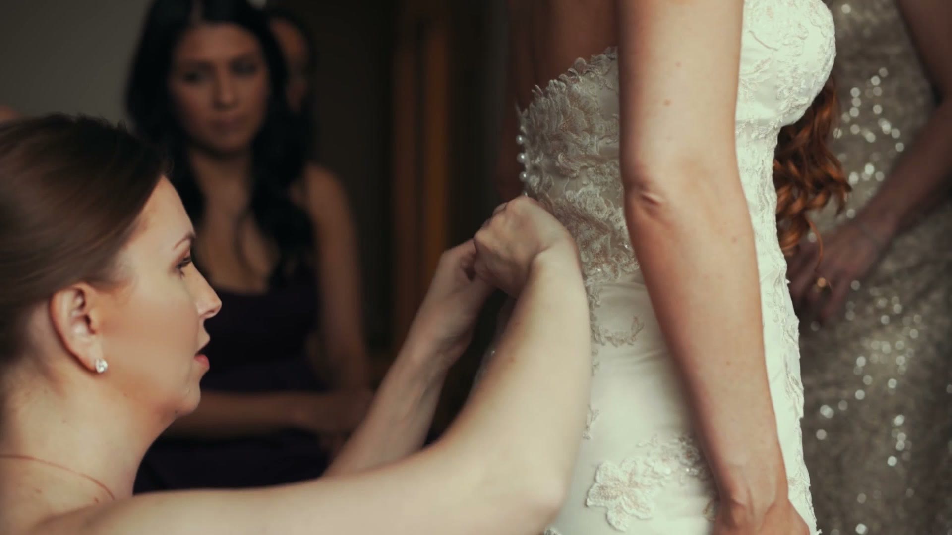 Bridesmaid lacing up the back of the wedding dress while another bridesmaid watches from the background
