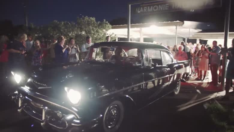 A vintage black car with its headlights on pulling away at night through a crowd of cheering guests under the Harmonie Hall sign