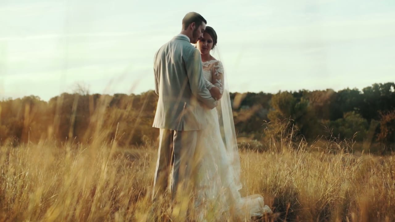 Bride and groom embracing close together in a golden field of tall grass, foreheads together, backlit by low sun