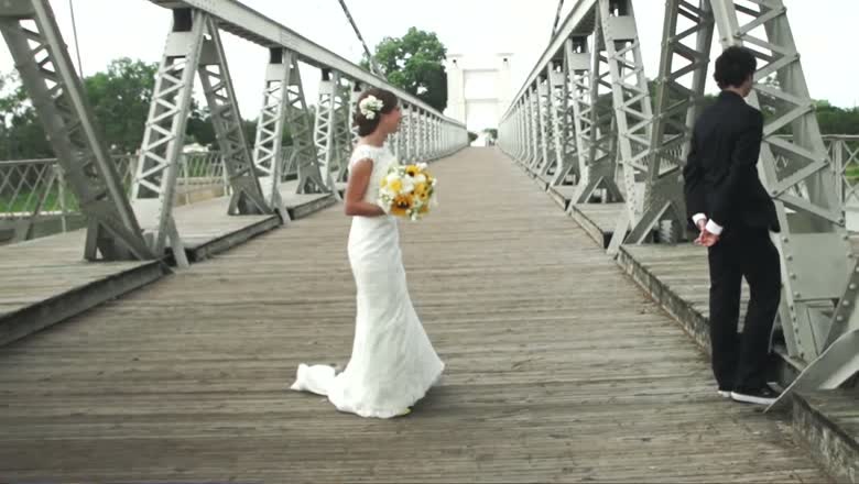 The bride standing on the wooden deck of the Waco suspension bridge holding a yellow bouquet, the groom facing away down the bridge