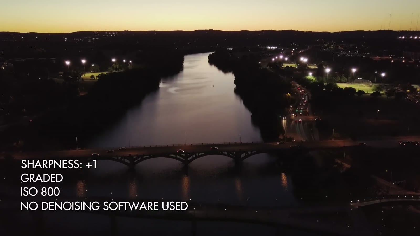 A dusk aerial of a river and lit bridges through a city, labelled Sharpness plus 1, graded, ISO 800, no denoising software used