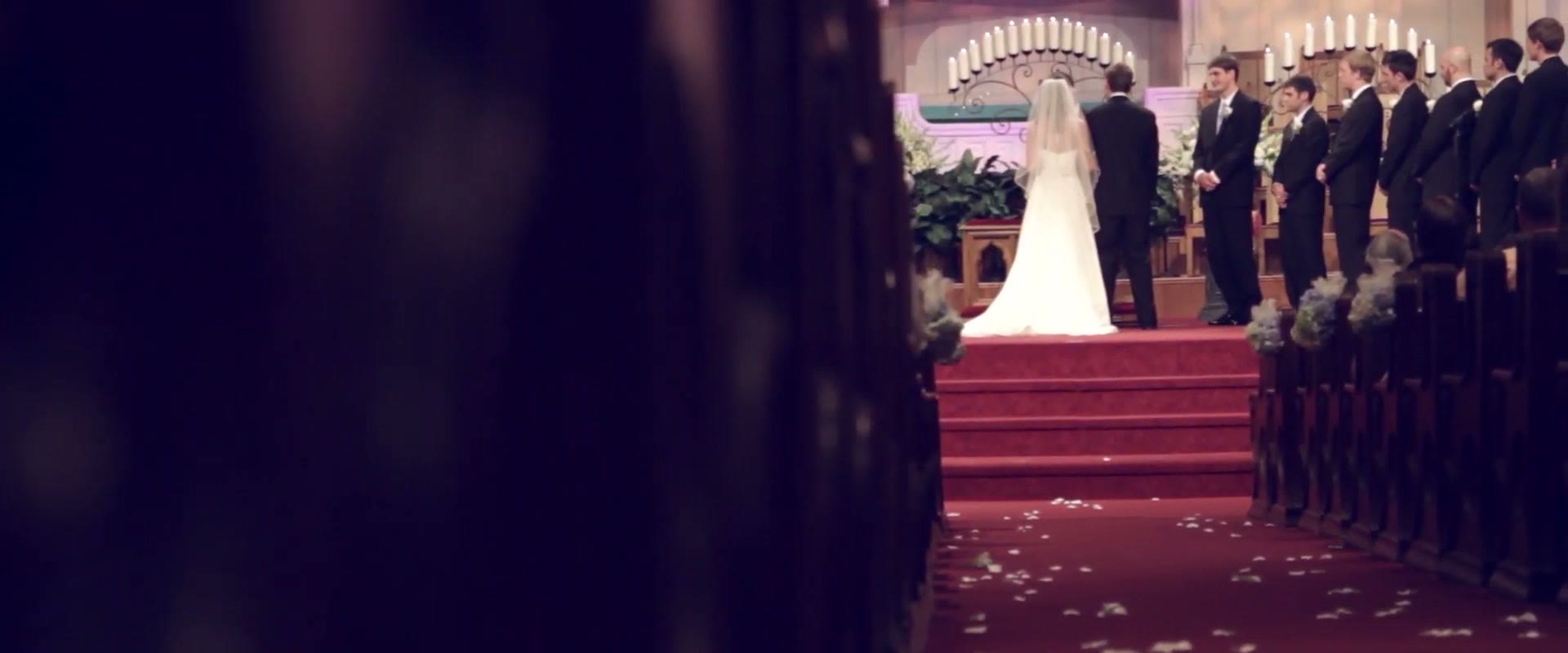 A bride standing at the top of the aisle of a large church, framed between dark wooden pews with petals scattered on the red carpet