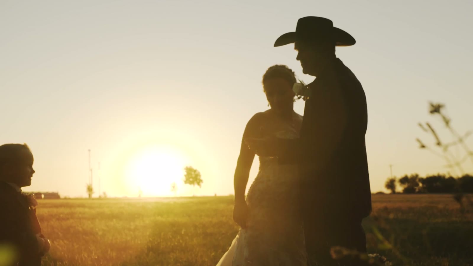 Silhouette of the bride and a cowboy-hatted groom against a setting sun, a child watching from the side