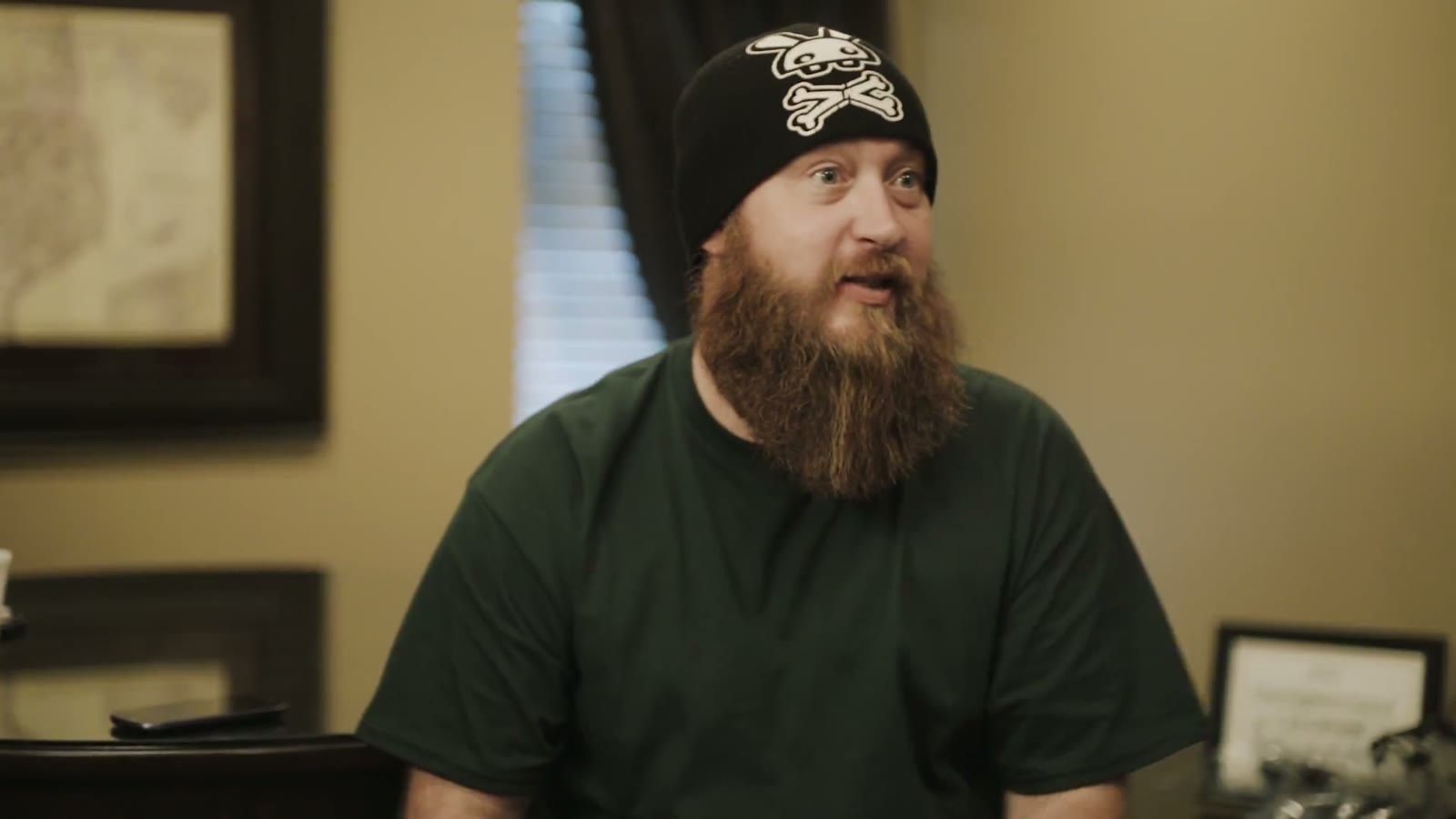 Groom in a black skull-and-crossbones beanie talking during an interview, framed portrait on the wall behind him