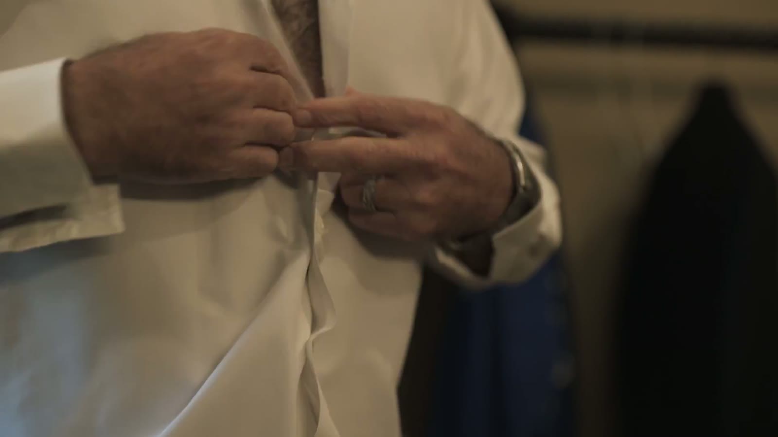 Close-up of hands buttoning a white dress shirt, a wedding ring visible on the ring finger