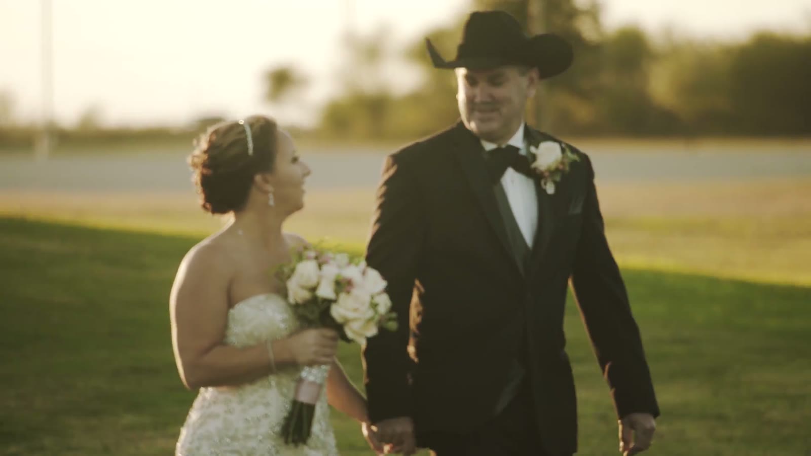 Bride and groom in a black cowboy hat walking hand in hand across a grassy field at golden hour