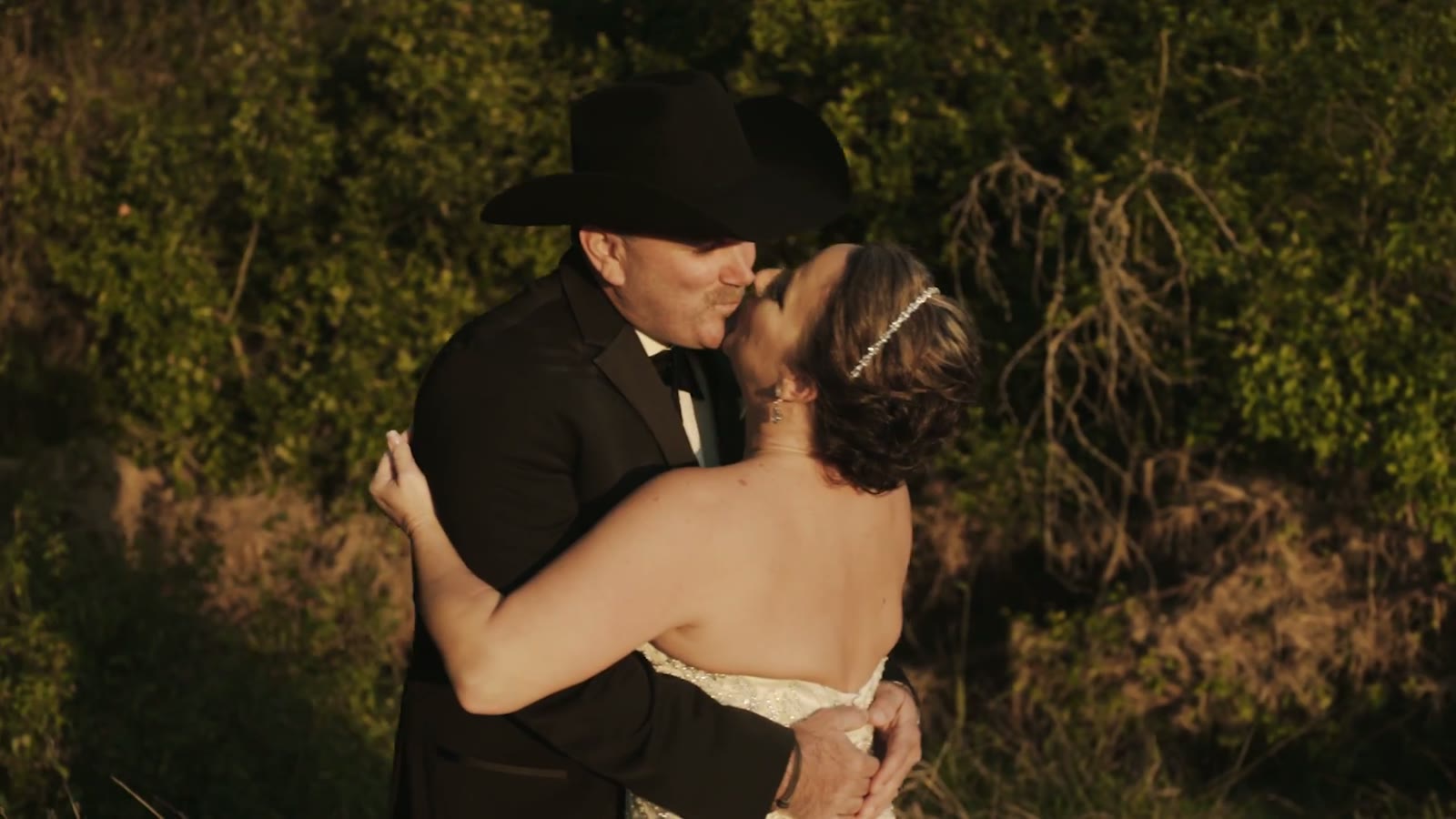 Groom in a black cowboy hat kissing the bride against a backdrop of brush at sunset