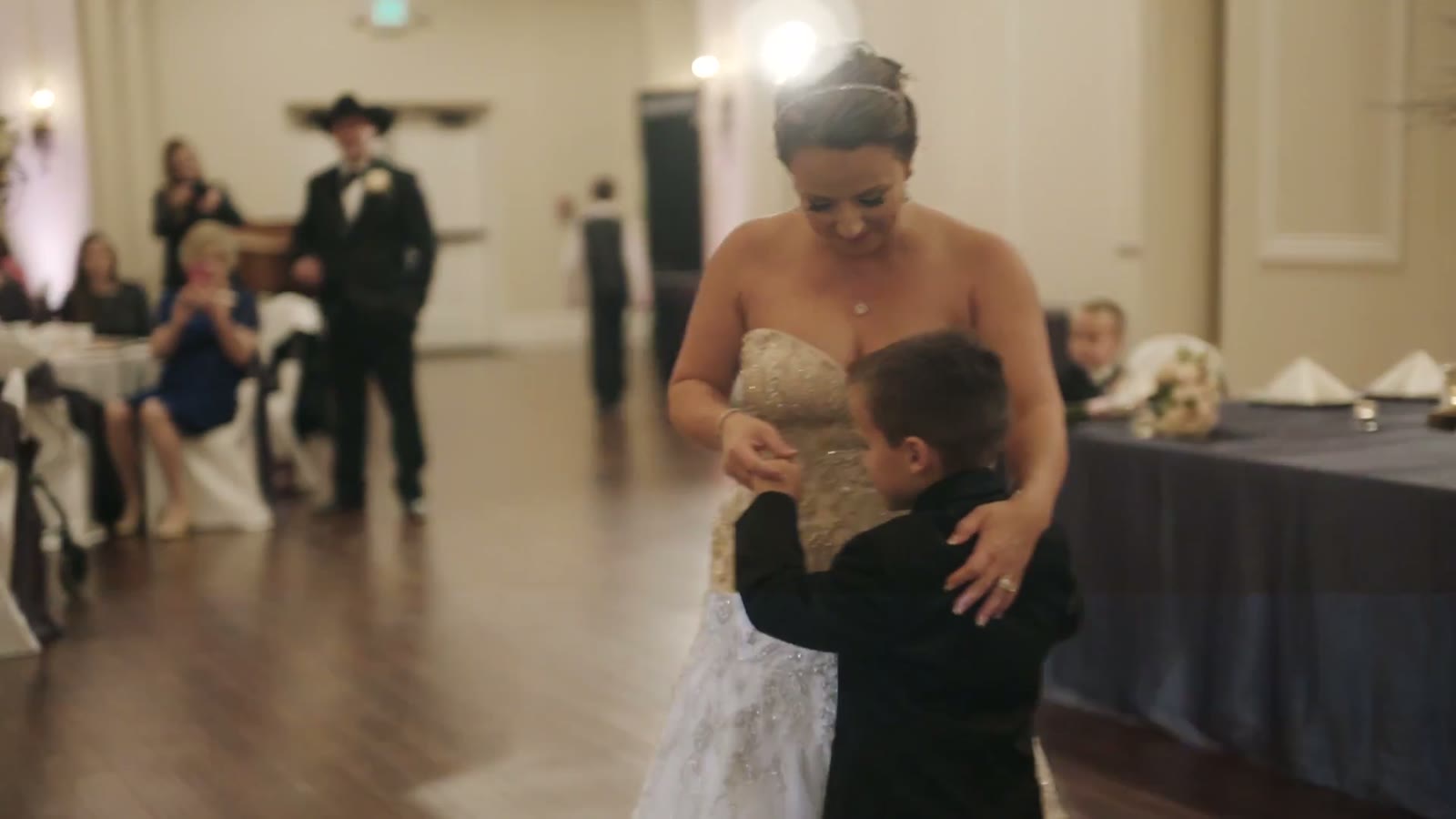 Bride dancing with a young boy in a suit on the reception dance floor, guests seated at tables behind them
