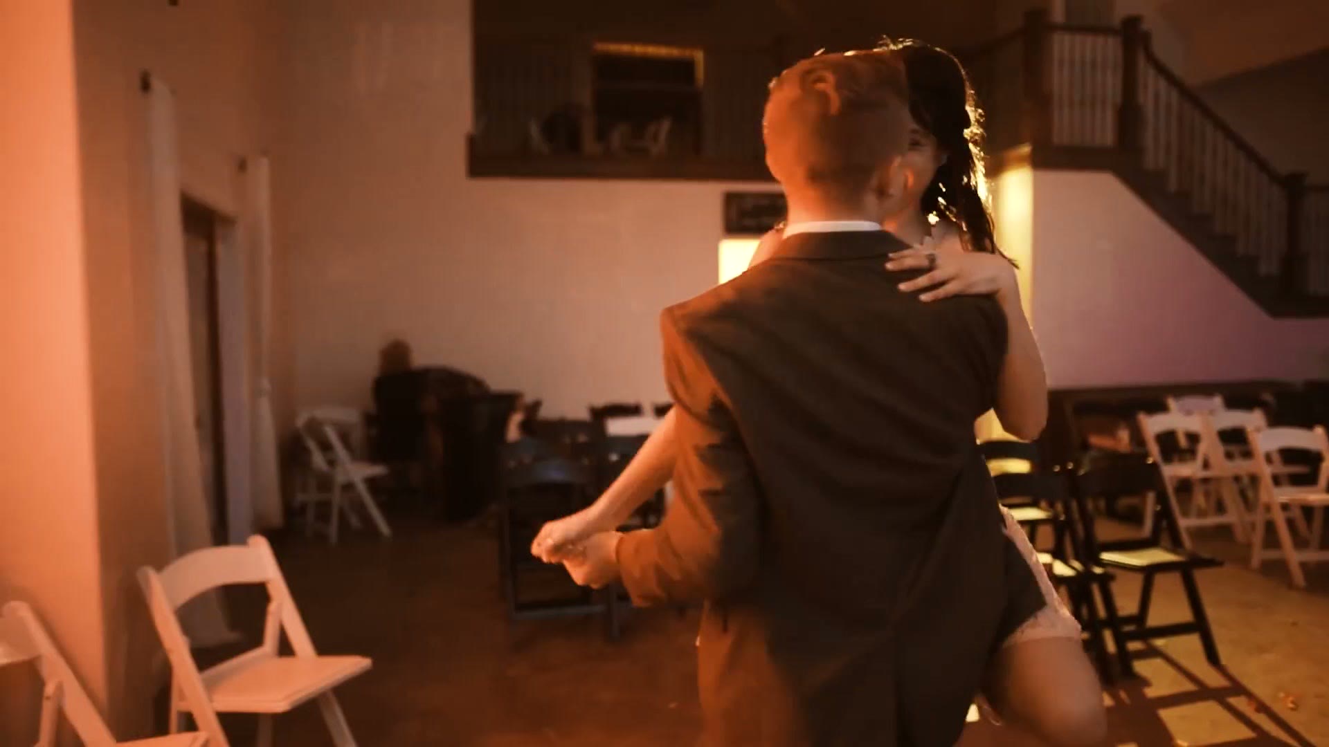 A bride and groom dancing alone together in an empty reception room lit warm and low, seen from behind