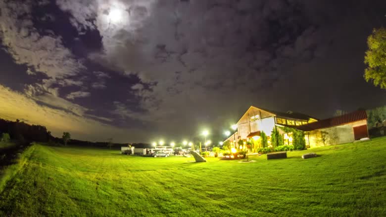 A night timelapse of the venue lit up across a wide lawn under a moonlit sky with clouds streaking past