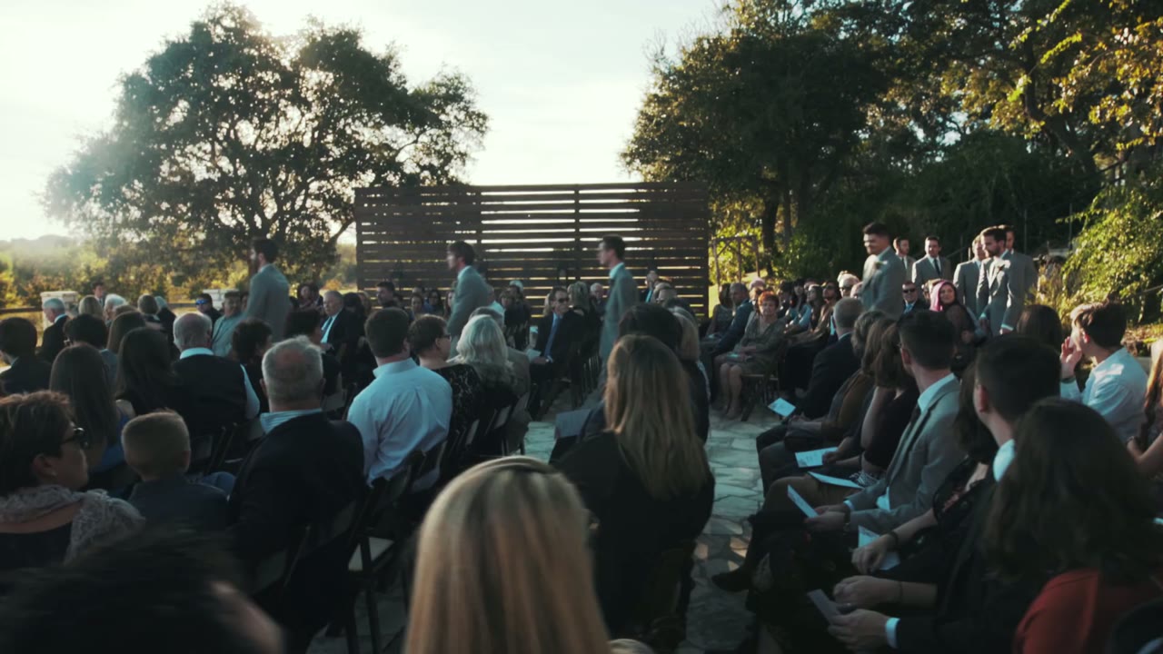 Outdoor hill country ceremony with rows of seated guests and the wedding party gathered under a wood-slat arch