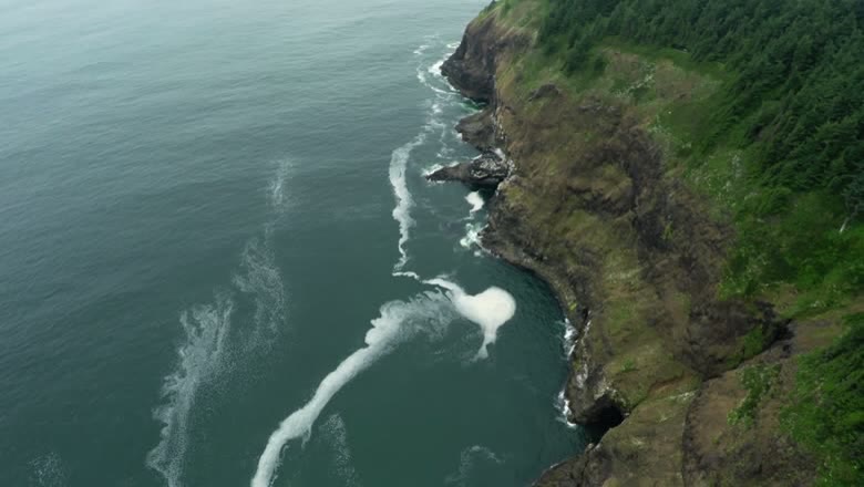 Looking straight down at white surf breaking along the base of a steep green headland dropping into deep teal water