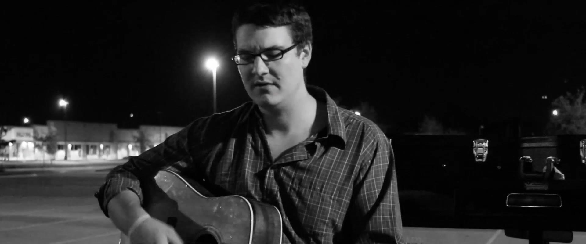 Black and white shot of the musician with eyes closed looking down in concentration while playing, a road case to his right