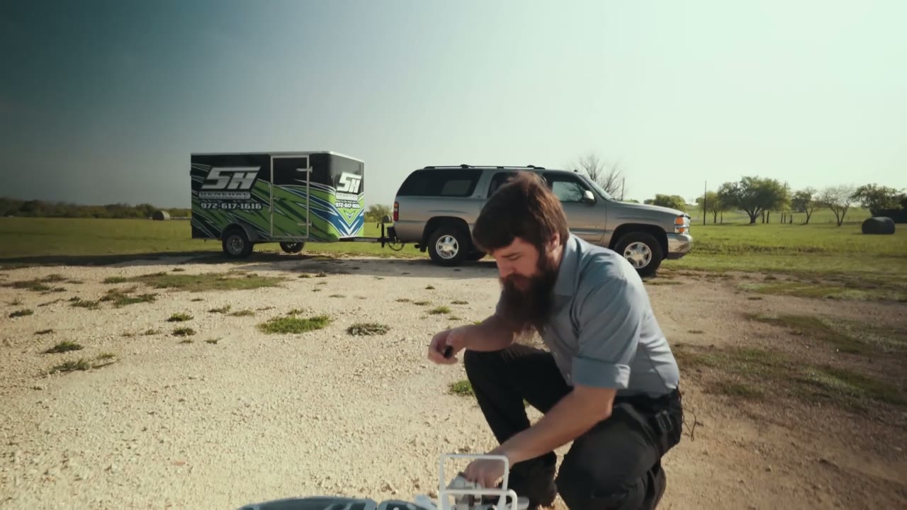 Matt kneeling to prep a DJI Phantom 4 drone in a field, an SH-branded trailer and SUV parked behind him