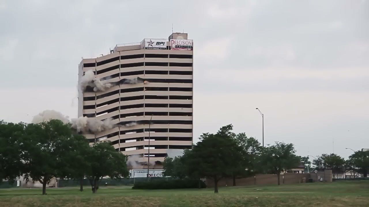 A tall concrete hotel tower still standing under an overcast sky, with demolition contractor banners hung across its upper floors and trees in the foreground