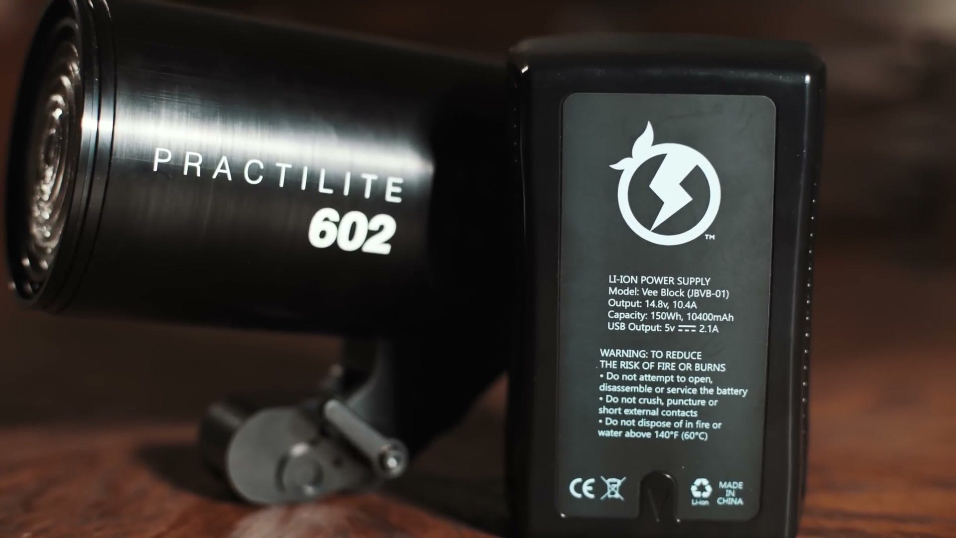 The Practilite 602 lying on a table beside a V-mount battery whose label reads 150 watt hours and 10400mAh