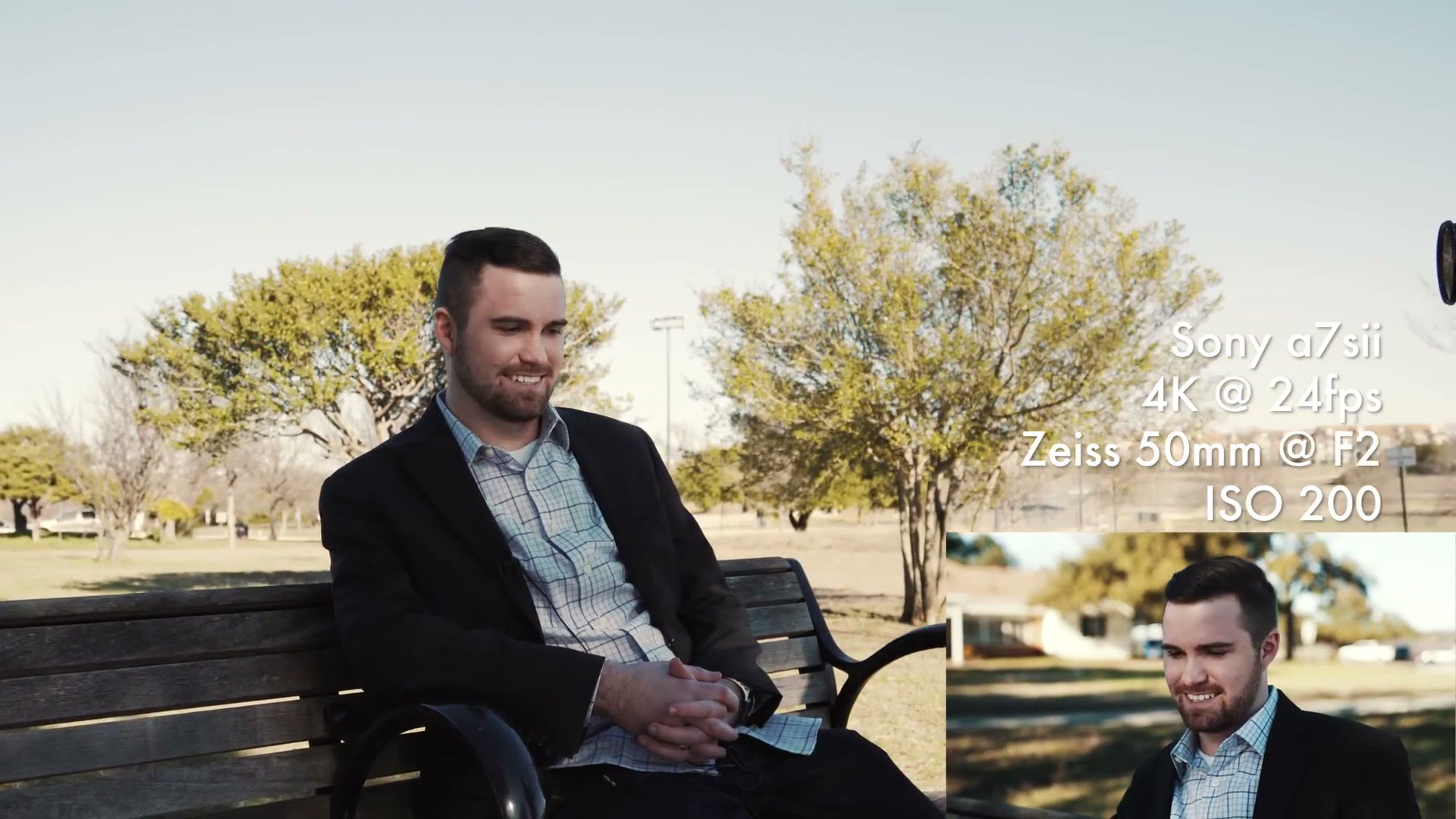 A man in a jacket and checked shirt sitting on a park bench for an interview, with on-screen text reading Sony a7sii, 4K at 24fps, Zeiss 50mm at F2, ISO 200.