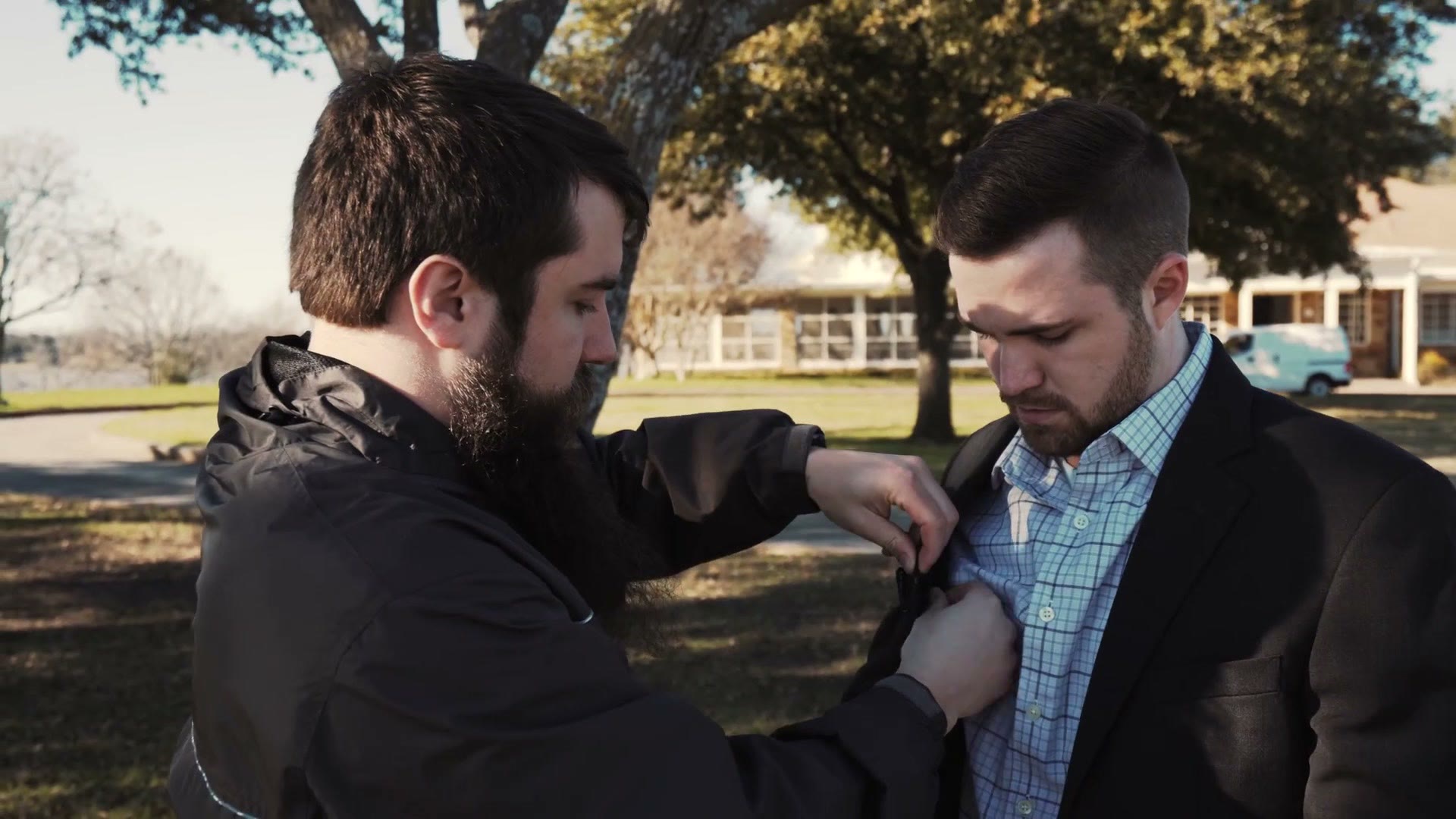 Matt clipping a lavalier microphone onto the shirt of the man who is about to propose, under a tree in a park.