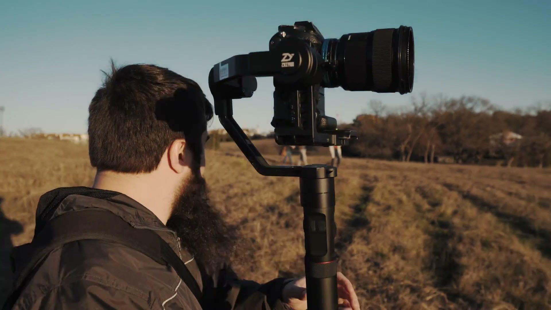 Close-up of a Zhiyun Crane 2 gimbal with a camera mounted on it, held up over Matt's shoulder in a field at golden hour.