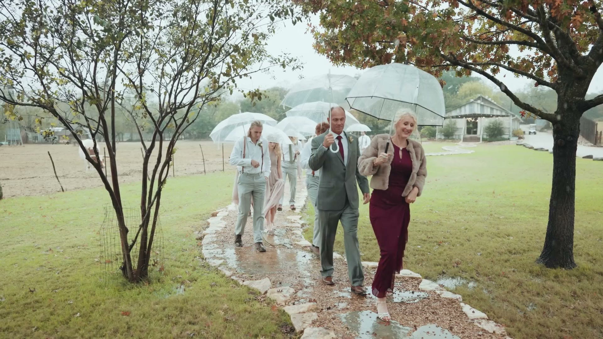 Wedding party walking along a wet stone path toward a chapel under clear bubble umbrellas in heavy rain
