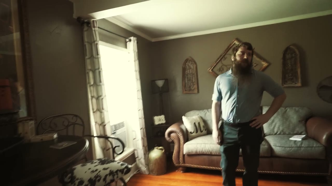 Matt standing hand on hip surveying the living room layout, couch and throw pillows arranged before filming